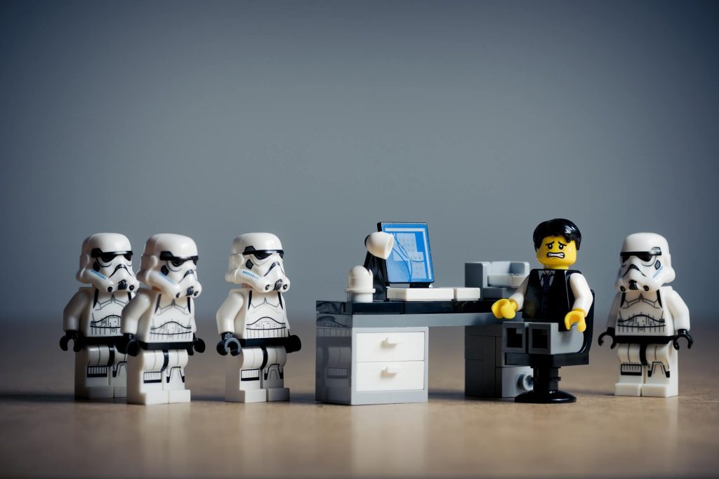 Les Stormtroopers LEGO projettent une présence intimidante autour d'un travailleur LEGO assis à un bureau, mettant en scène une représentation ludique de la dynamique du pouvoir dans un monde LEGO.